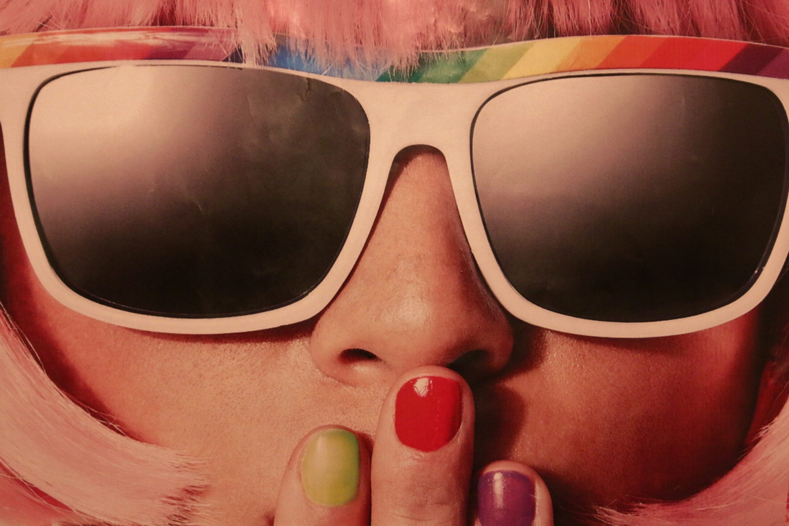 Nahaufnahme einer Person mit pinken Haaren, großen Sonnenbrillen und bunt lackierten Fingernägeln, Regenbogenband auf der Brille