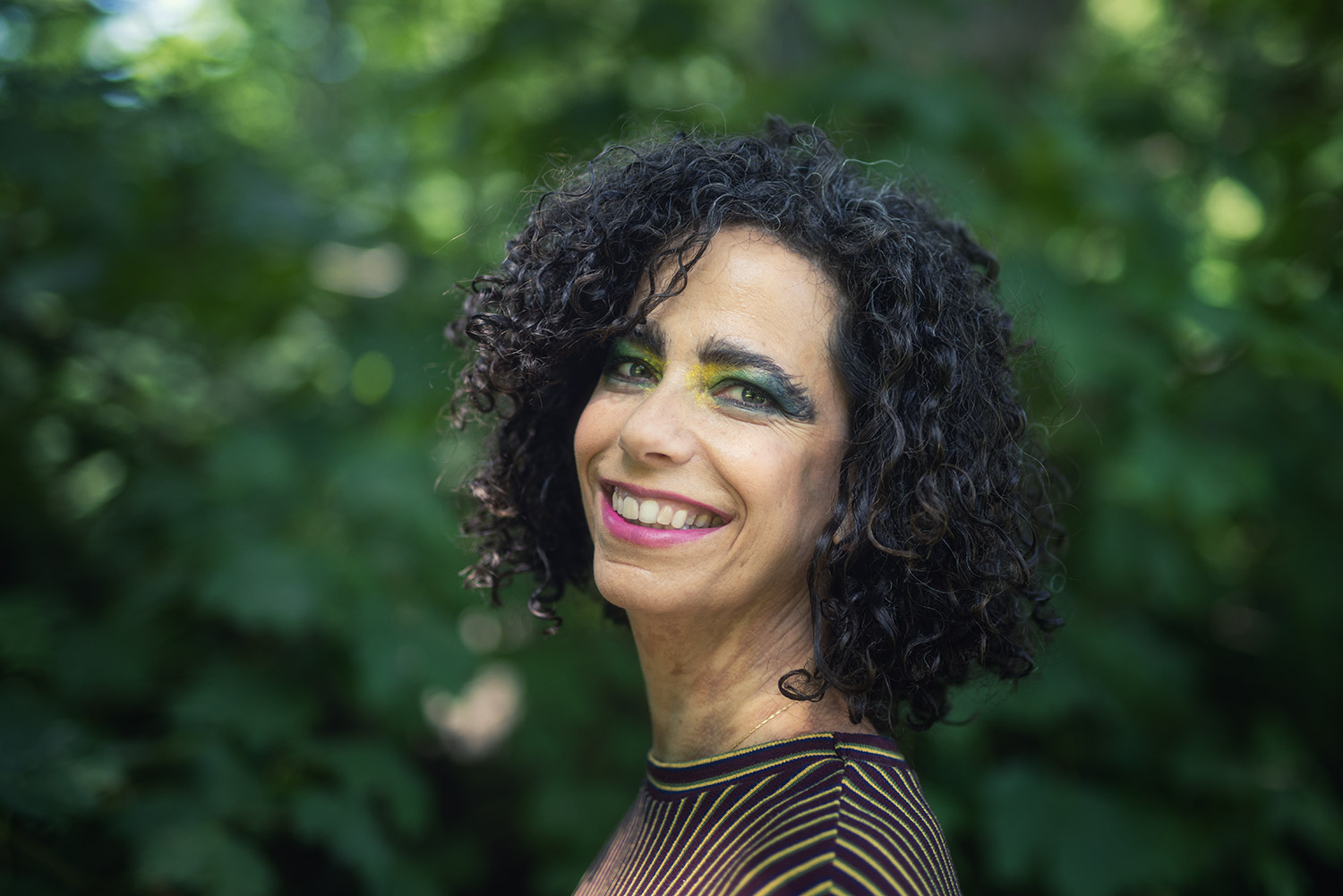 Person mit lockigem, dunklem Haar vor grünem, unscharfem Hintergrund aus Blättern und Bäumen. Die Person trägt ein Oberteil mit feinen, horizontalen Streifen in dunklen und hellen Tönen.