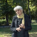 Person mit grauem Haar steht im Park vor grünen Bäumen und Rasenfläche. Die Person trägt einen dunklen Mantel, einen gestrickten Pullover und einen hellen Schal.