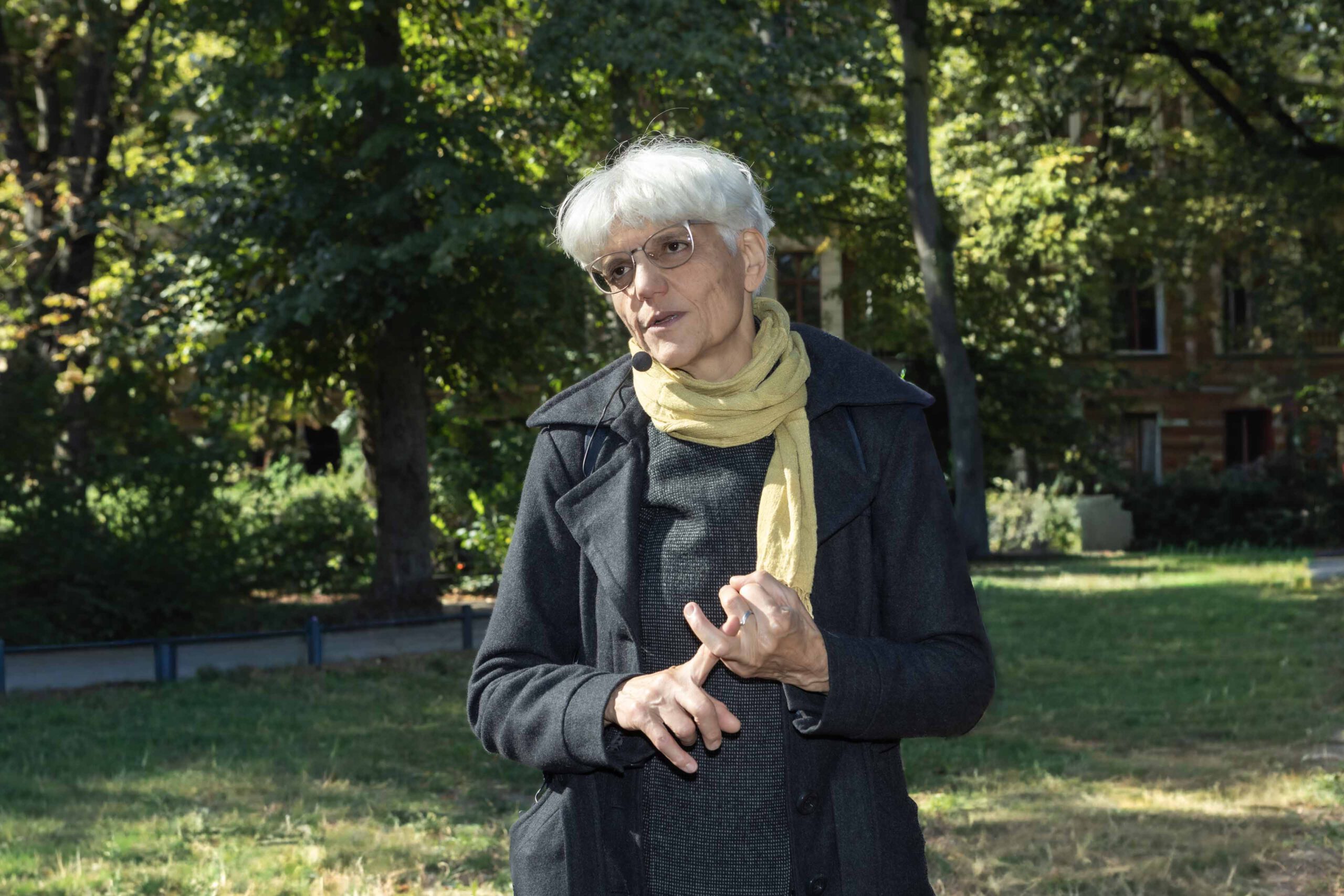 Person mit grauem Haar steht im Park vor grünen Bäumen und Rasenfläche. Die Person trägt einen dunklen Mantel, einen gestrickten Pullover und einen hellen Schal.