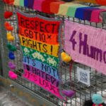 Bunter Zaun mit gehäkelten Botschaften für LGBTIQ-Rechte, darunter ‚RESPECT LGBTIQ RIGHTS! IT'S NOT HATE, IT'S LOVE‘ auf Regenbogenhintergrund und ‚4 Human Rights‘ auf pinkem Stoff. Kleine Herzdekorationen und ein QR-Code sind sichtbar.