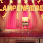 Bühne mit rotem Vorhang, einem Stuhl im Scheinwerferlicht, Klavier links und Stehlampe rechts; darüber der Schriftzug ‚LAMPENFIEBER‘ mit gelben Blitzen.
