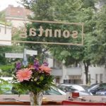 Blick aus einem Café mit Blumen auf dem Tisch, Schriftzug ‚sonntags club‘ am Fenster, draußen Straße mit Autos und Straßenschild ‚Erich-Weinert-Straße‘
