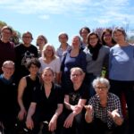 Gruppenfoto von fünfzehn Personen auf einer Terrasse im Freien, mit Bäumen und blauem Himmel im Hintergrund.