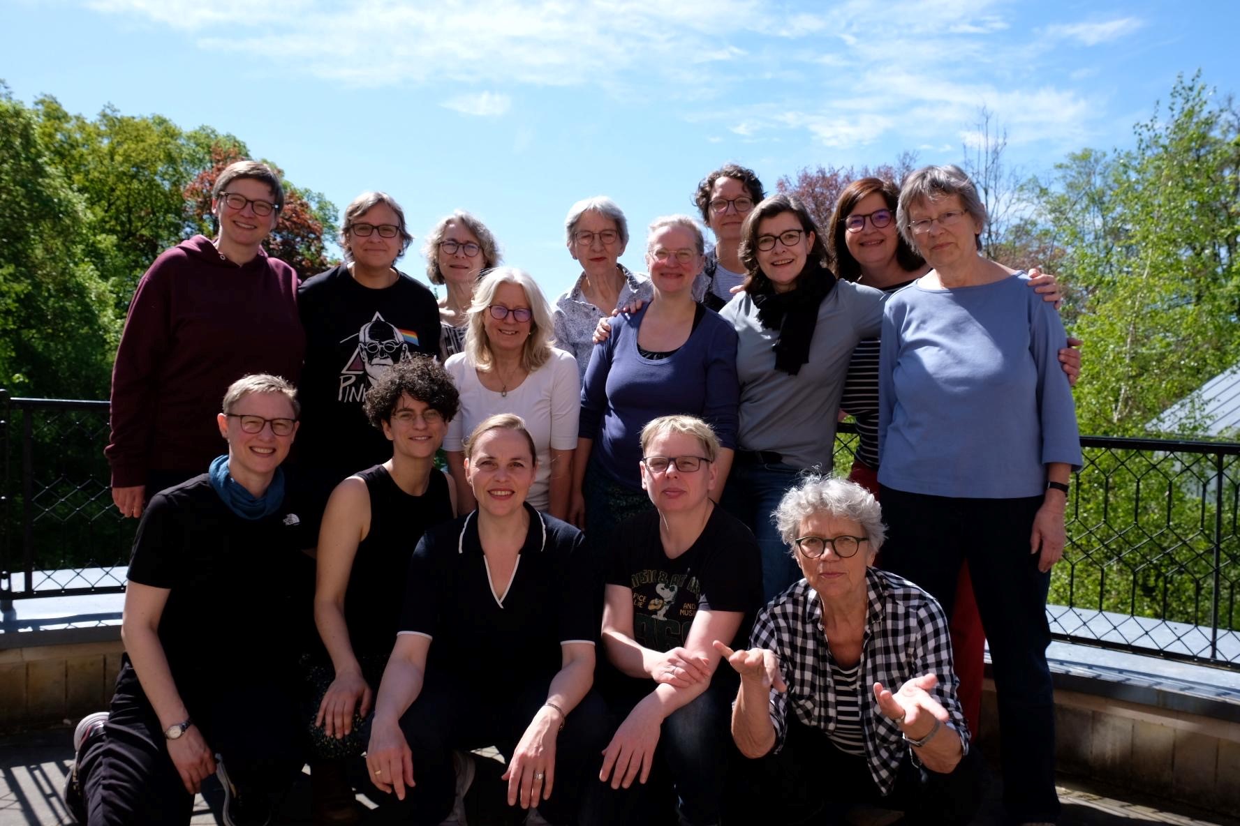 Gruppenfoto von fünfzehn Personen auf einer Terrasse im Freien, mit Bäumen und blauem Himmel im Hintergrund.
